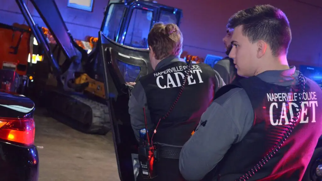 Two Naperville police cadets with vests on and using shield walking through room with large equipment in competition