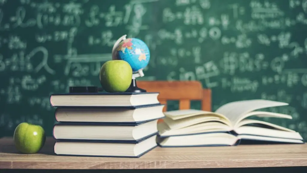 Stack of books sitting on school desk along with 2 apples and a small globe with chalkboard with equations written on it in background.