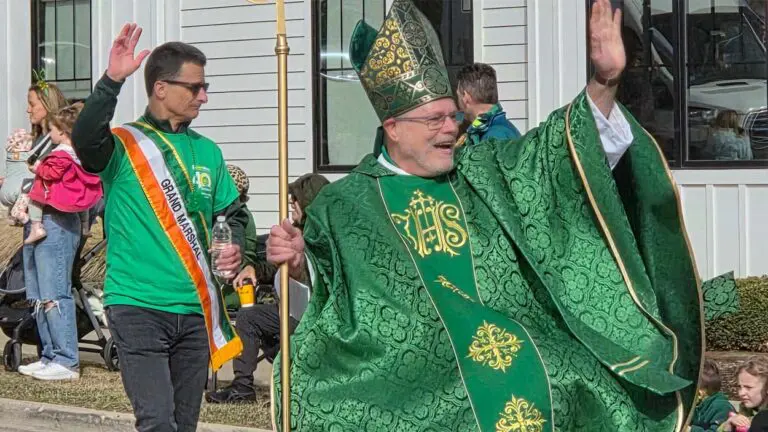 A man dressed as St. Patrick waves to people while the parade grand marshal is behind him, also waving.
