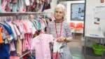 A woman holds a pink toddler outfit in a donation room full of children's clothes