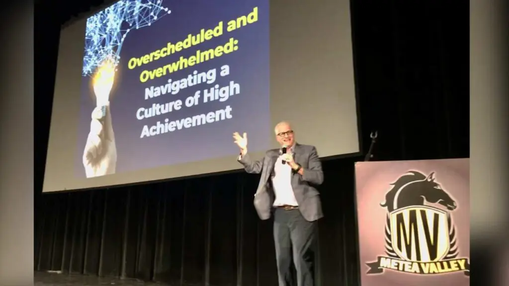 Doug Bolton on stage at Mental Health Symposium with screen in background with slide about being overscheduled and overwhelmed