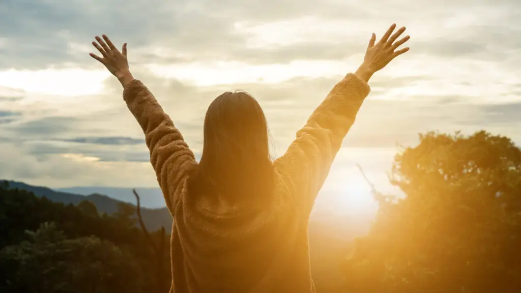 A woman faces the sunset with her arms raised and hands open.