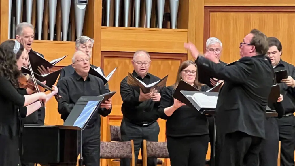 A group of singers dressed in all black hold binders while the conductor directs them.