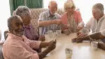 A group of senior citizens are gathered around a table playing cards while one smiles a the camera.