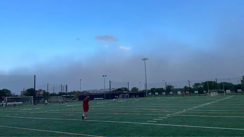 Man in red outfit walks across soccer field with large dust storm cloud visible in distance