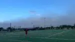 Man in red outfit walks across soccer field with large dust storm cloud visible in distance