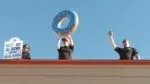 3 police personnel atop a Dunkin' rooftop, one holding a sign, another holding an inflatable donut