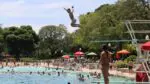 A view of a large pool with a young boy jumping off the high dive and people in the water in the distance.