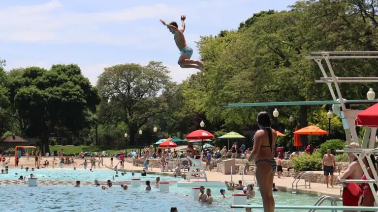 A view of a large pool with a young boy jumping off the high dive and people in the water in the distance.