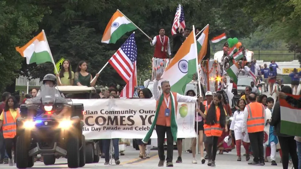 A group of people participating in the India Day celebration walk in the parade while waving Indian and American flags to onlookers.
