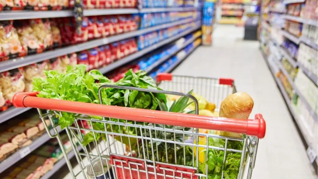 Grocery cart with some produce items in top of cart with view down the aisle of a grocery store for grocery sales tax story