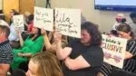 Speakers at D203 board meeting holding signs mostly against transgender athlete participation in girls sports, with one person holding sign on the opposite side of issue