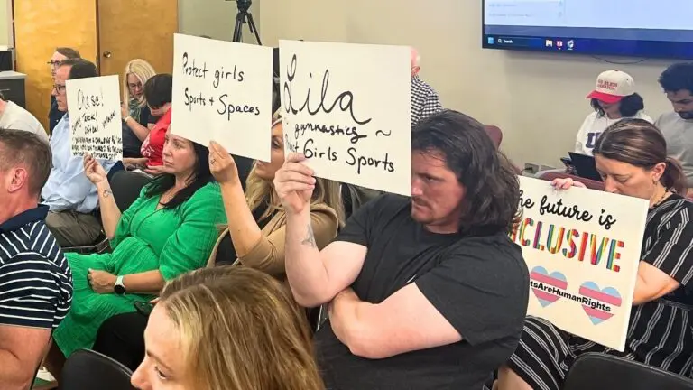 Speakers at D203 board meeting holding signs mostly against transgender athlete participation in girls sports, with one person holding sign on the opposite side of issue
