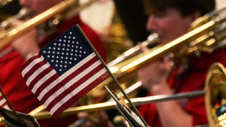 An American flag is in focus, with a trombone player behind it, out of focus.