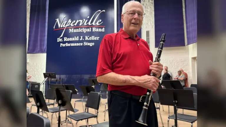 Dick Wheeler holding a clarinet, standing in front ot the Naperville Municipal Band stage sign