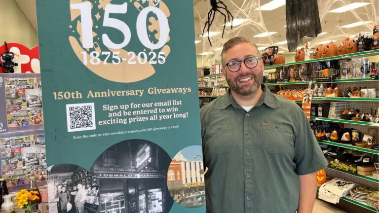Sixth-generation owner of Oswald's Pharmacy Alex Anderson smiles for a picture with a sign promoting their 150th anniversary.