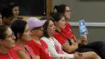 People in red shirts supporting teachers at District 203 sit in chairs at D203 board meeting one holding sign