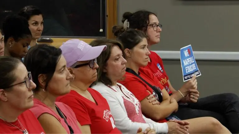 People in red shirts supporting teachers at District 203 sit in chairs at D203 board meeting one holding sign