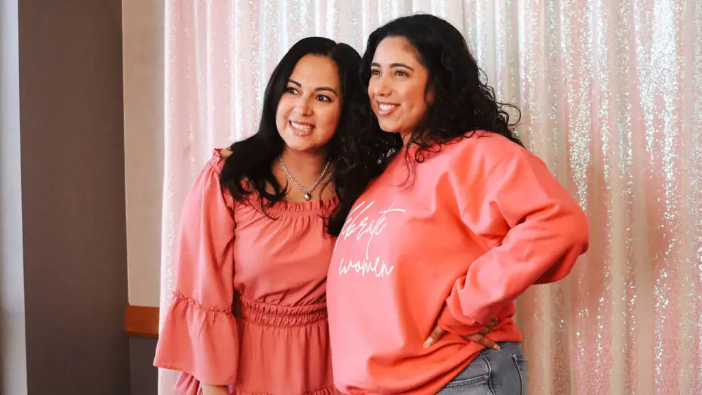 Two young women wear pink and smile in front of a glitter curtain backdrop.