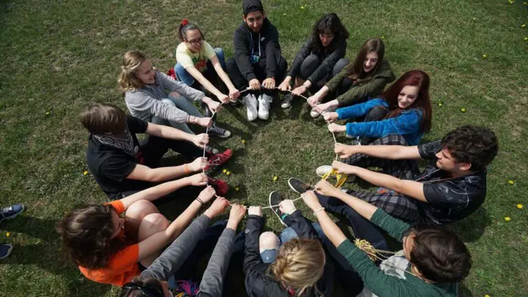 A group of high school students sit in a circle and hold their hands in a ring in front of them.