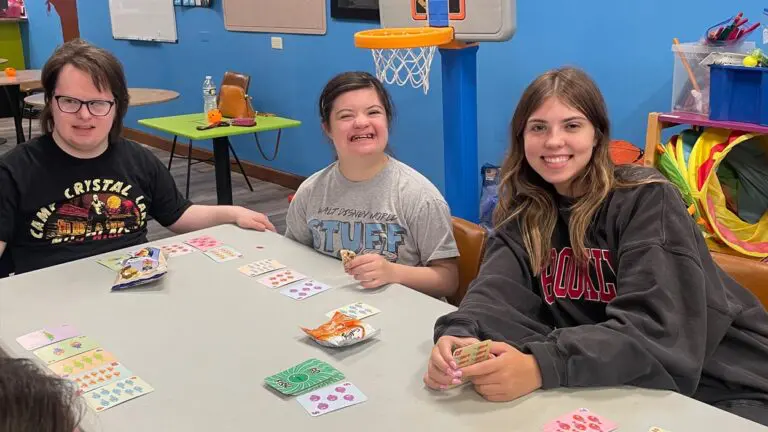 Two GiGi's Playhouse participants play cards with a volunteer.