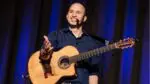 Jim Perona, while holding a guitar, smiles at a crowd at one of his keynote concerts.