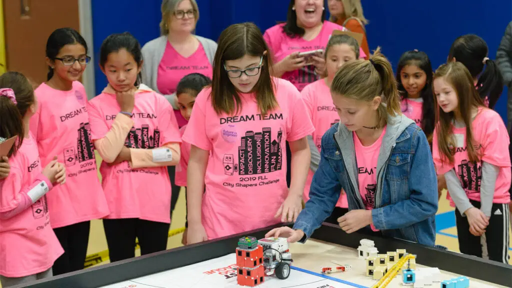 A group of elementary aged girls stand around a table with robotic Legos. They are wearing pink t-shirts that say dream team.