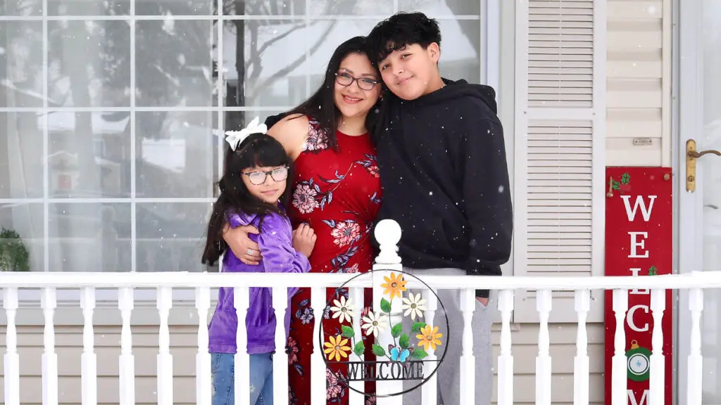 A family of three, mom, daughter, and son, stand on their new porch adorned with a welcome sign.