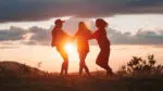 Three girls, in silhouette with the sun behind them, jump together.