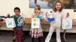 Three children hold up handmade snowman decorations and festive bags.
