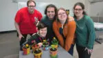 A group of five individuals smile in front of hand-painted pots with flowers planted in them.