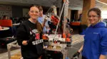 Three students working on their robot with FIRST Robotics, a male student in the background, and two female students smile at the camera in the foreground.