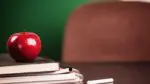 books and notebooks stacked with an apple on top on teacher desk with an empty chair in background