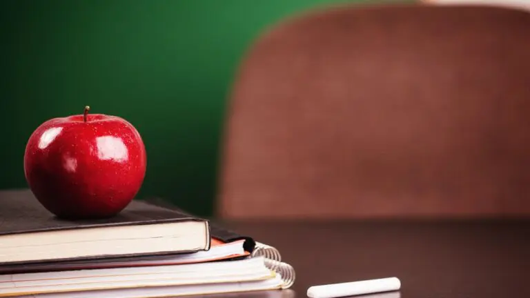 books and notebooks stacked with an apple on top on teacher desk with an empty chair in background