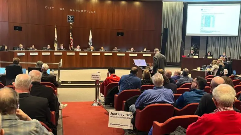 Wide shot of Naperville city council meeting with view of the dais and some audience members along nwith a sign