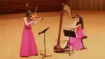 Two girls in pink dresses perform at an OPUS concert. The left plays violin and the right plays the harp.