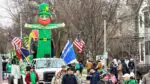A 2026 St. Patrick's Day Parade entry with a group walking in front of a truck with a large, blow up leprechaun in the bed.