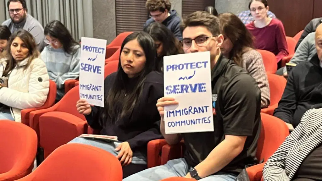 two people at Naperville City Council meeting holding up Protect and Serve immigrant communities signs