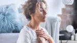 A woman with closed eyes sits on the floor with her hands clasped against her chest with a peaceful look on her face.