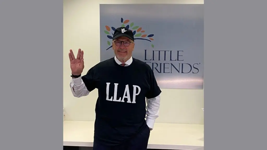 Little Friends CEO Mike Briggs standing in front of organization sign making the Vulcan greeting sign with his hand