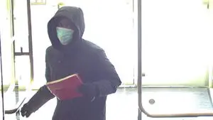 A man is seen walking while carrying a red folder. He is wearing a black hoodie, a surgical face mask and black gloves.