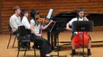 A chamber music ensemble on the Wentz Concert Hall Stage at North Central College; a violin player, a piano player, and a cello player, while an instructor sits by the piano, ready to turn pages.