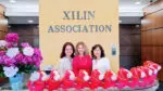 Three women stand in front of a sign that reads "Xilin Association" and on the desk in front of them are Fire Horse plushies, the symbol of the 2026 Chinese Zodiac.