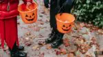 Two children holding plastic trick-or-treat buckets, leaves are on the ground