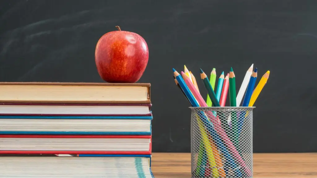 Stack of books with apple on top sitting on desk with container of colored pencils next to it and chalkboard in background