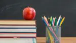 Stack of books with apple on top sitting on desk with container of colored pencils next to it and chalkboard in background