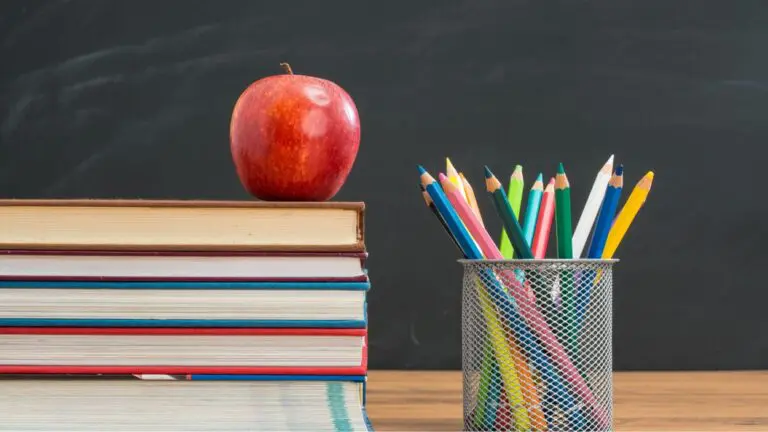 Stack of books with apple on top sitting on desk with container of colored pencils next to it and chalkboard in background