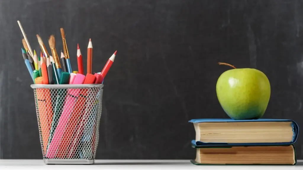 Stack of books with an apple and pencil cup holder in front of a blackbaord