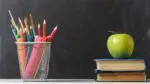 Stack of books with an apple and pencil cup holder in front of a blackbaord