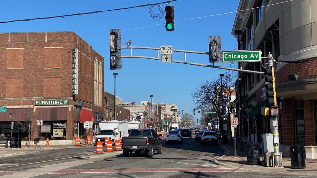 Downtown Naperville street with buildings, traffic lights and cars in view, as well as orange traffic barrels where streetscape work will happen